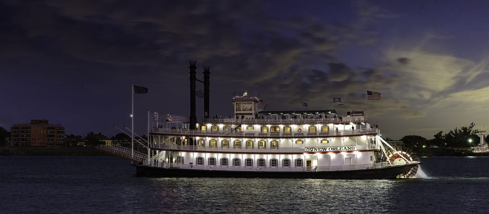 A three-deck paddlewheel riverboat with its decks lit up, cruising the Mississippi River at New Orleans at dusk